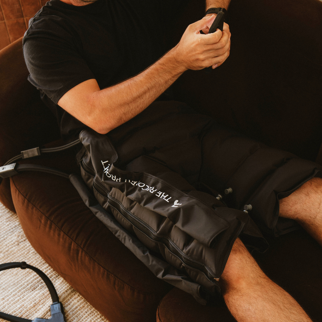 Man on a couch wearing the Lower Body Compression hip attachment, holding the control unit
